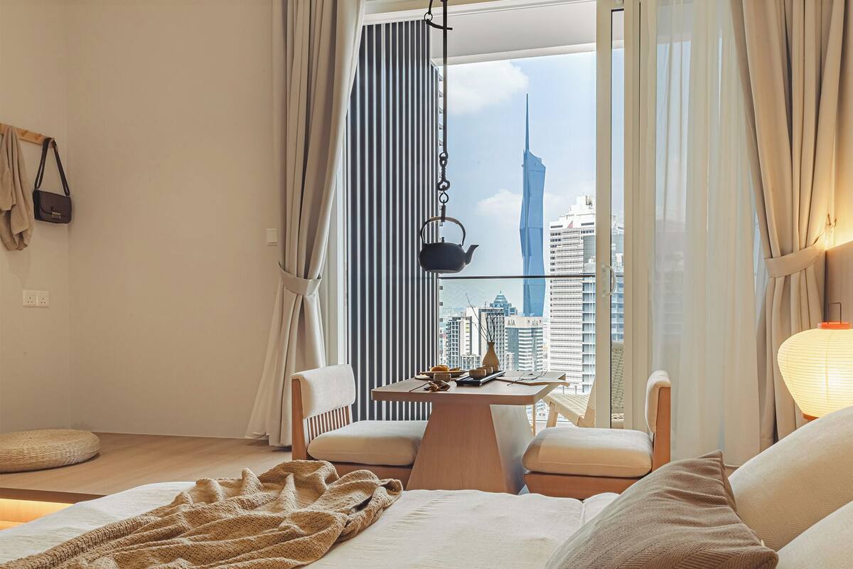 Une chambre d’hôtel moderne dotée d’une grande fenêtre, d’un lit, d’une petite table avec une bouilloire et offrant une vue sur le paysage urbain.