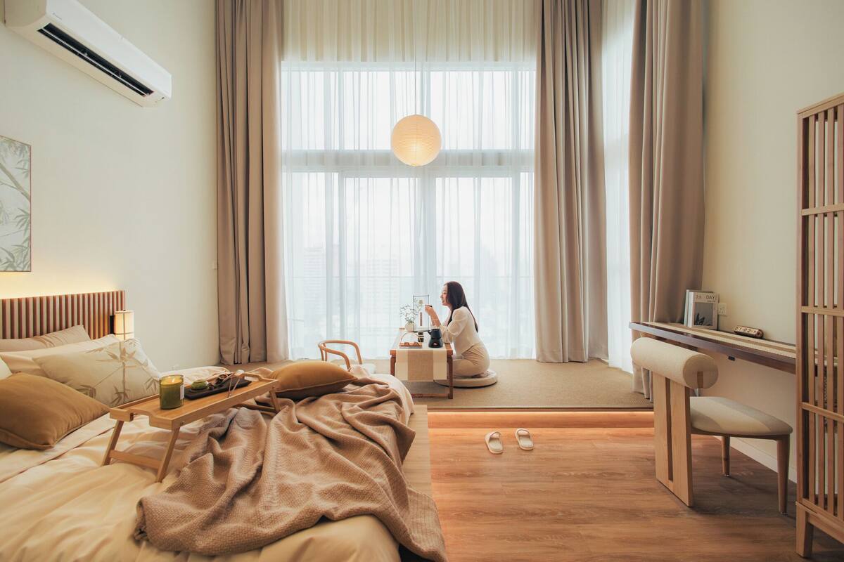 Une chambre moderne avec une grande fenêtre, une table de chevet avec une lampe, un lit avec une couverture, un petit bureau avec une chaise et un plancher en bois.