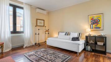 Ett modernt hotellrum med en vit soffa, en mönstrad matta, ett element, ett fönster med gardiner, en inramad tavla, en lampa och en bokhylla.