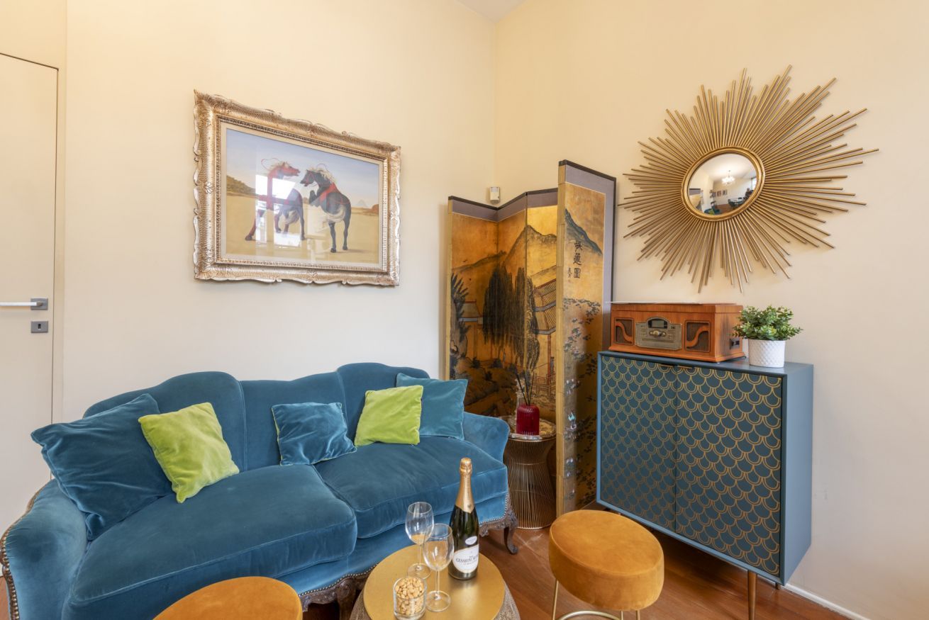 Ein Zimmer mit einem blauen Sofa, einem kleinen runden Tisch mit einer Flasche und Gläsern, einem dekorativen Spiegel und einer Kommode mit einem gemusterten Design.