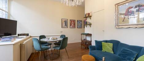 Una sala de estar con un sofá azul, una araña, una mesa de comedor con sillas y un televisor sobre un mueble.