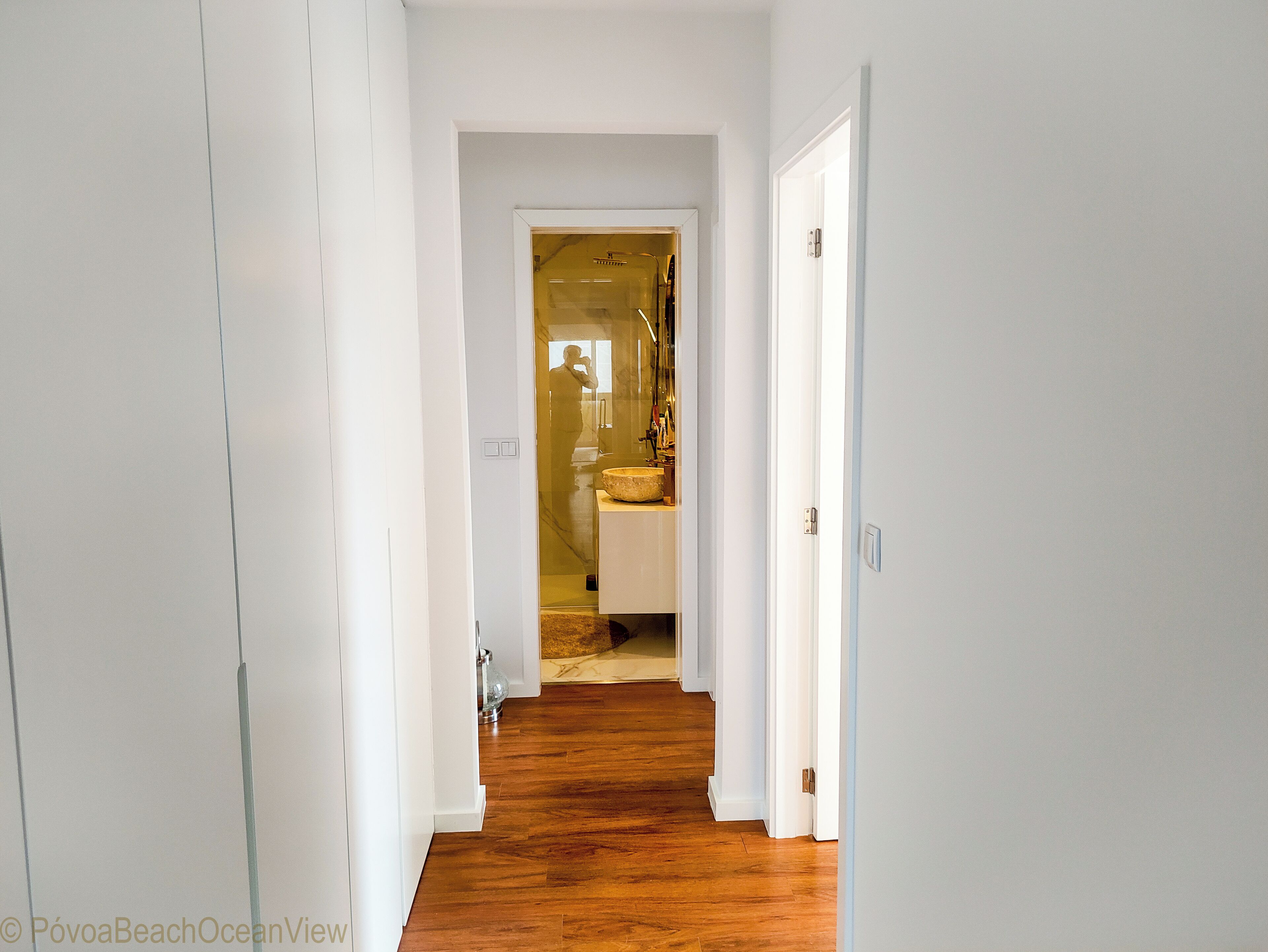 A hallway with wooden flooring leading to a bathroom with a sink and mirror.