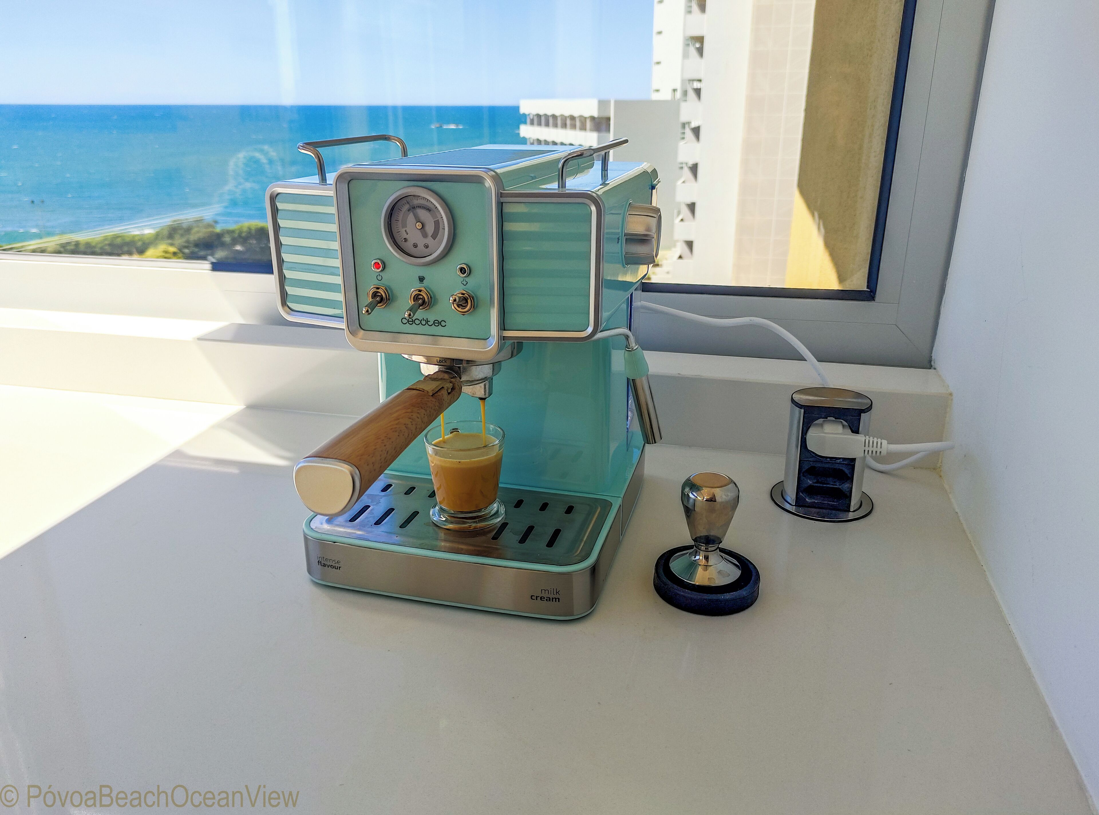 A vintage espresso machine on a balcony with a view of the ocean and buildings in the background.