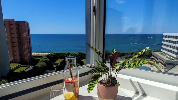 Un balcon offrant une vue dégagée sur l’océan et les bâtiments, une bouteille en verre contenant une boisson et une plante en pot.