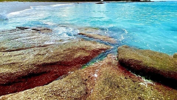 Ein Strand mit klarem türkisblauem Wasser, einem felsigen Vordergrund und einer fernen Küstenlinie unter einem teils bewölkten Himmel.