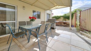 An outdoor patio with a glass table and chairs, a barbecue grill, and a covered area with a metal roof.