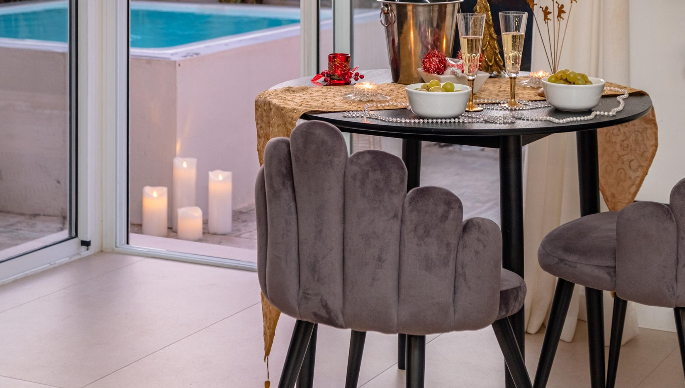 Ein Tisch ist mit Kerzen, einer Traube und einem Glas Champagner gedeckt, im Hintergrund ist ein Pool durch eine Glastür sichtbar.