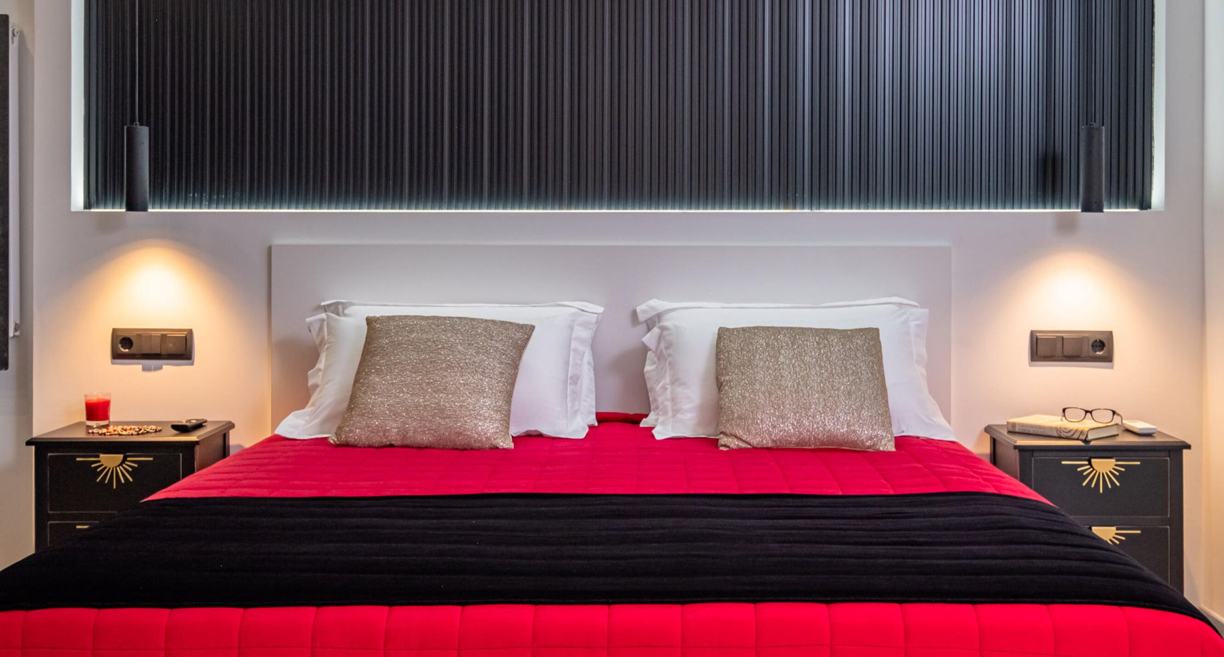 Un lit recouvert d’une couvre-lit rouge et noire, avec deux oreillers blancs et deux oreillers dorés. Il y a deux tables de chevet avec un revêtement noir et des détails dorés.