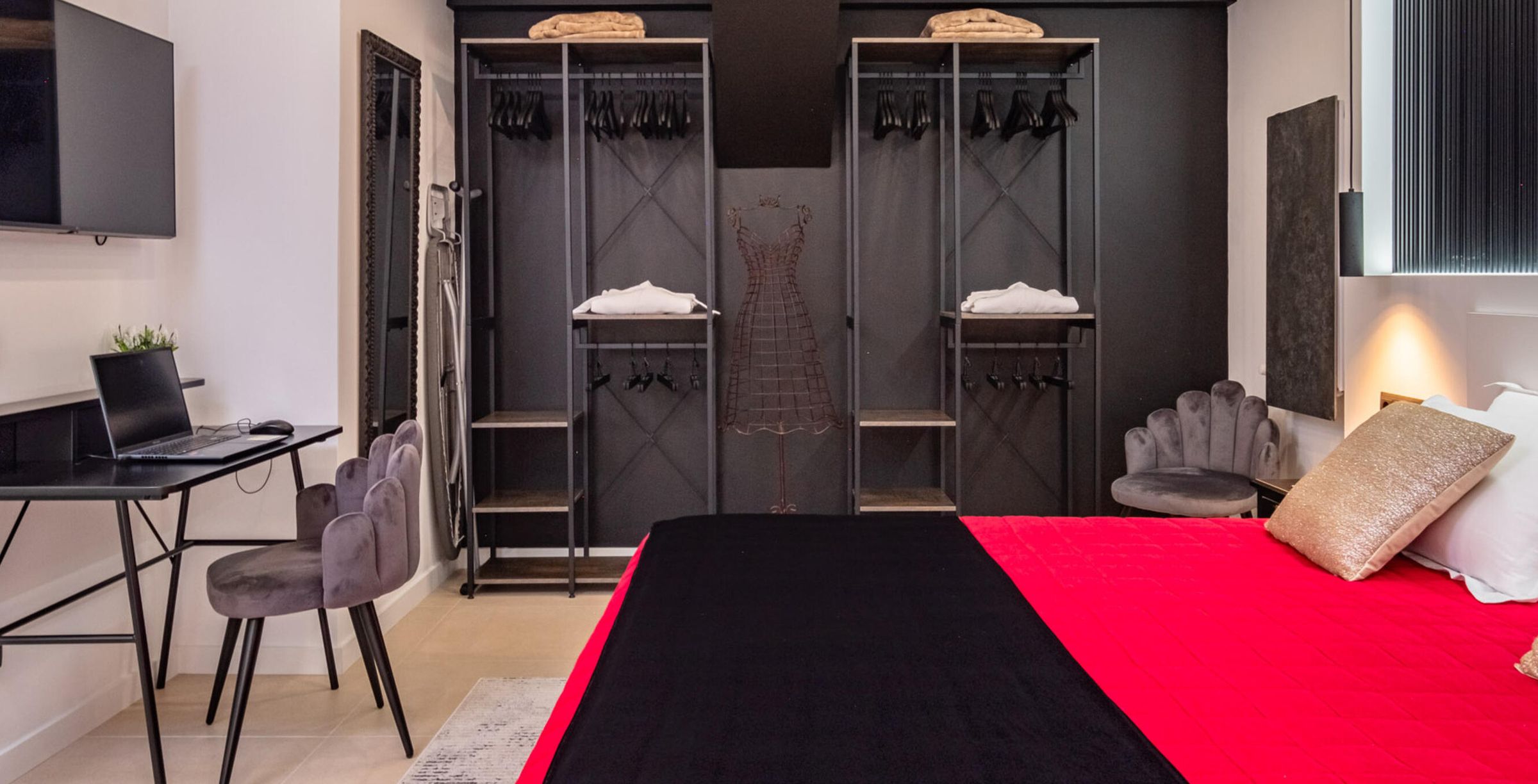 A bedroom with a bed, a desk with a laptop, a chair, a wardrobe with hanging clothes, and a nightstand with a lamp.