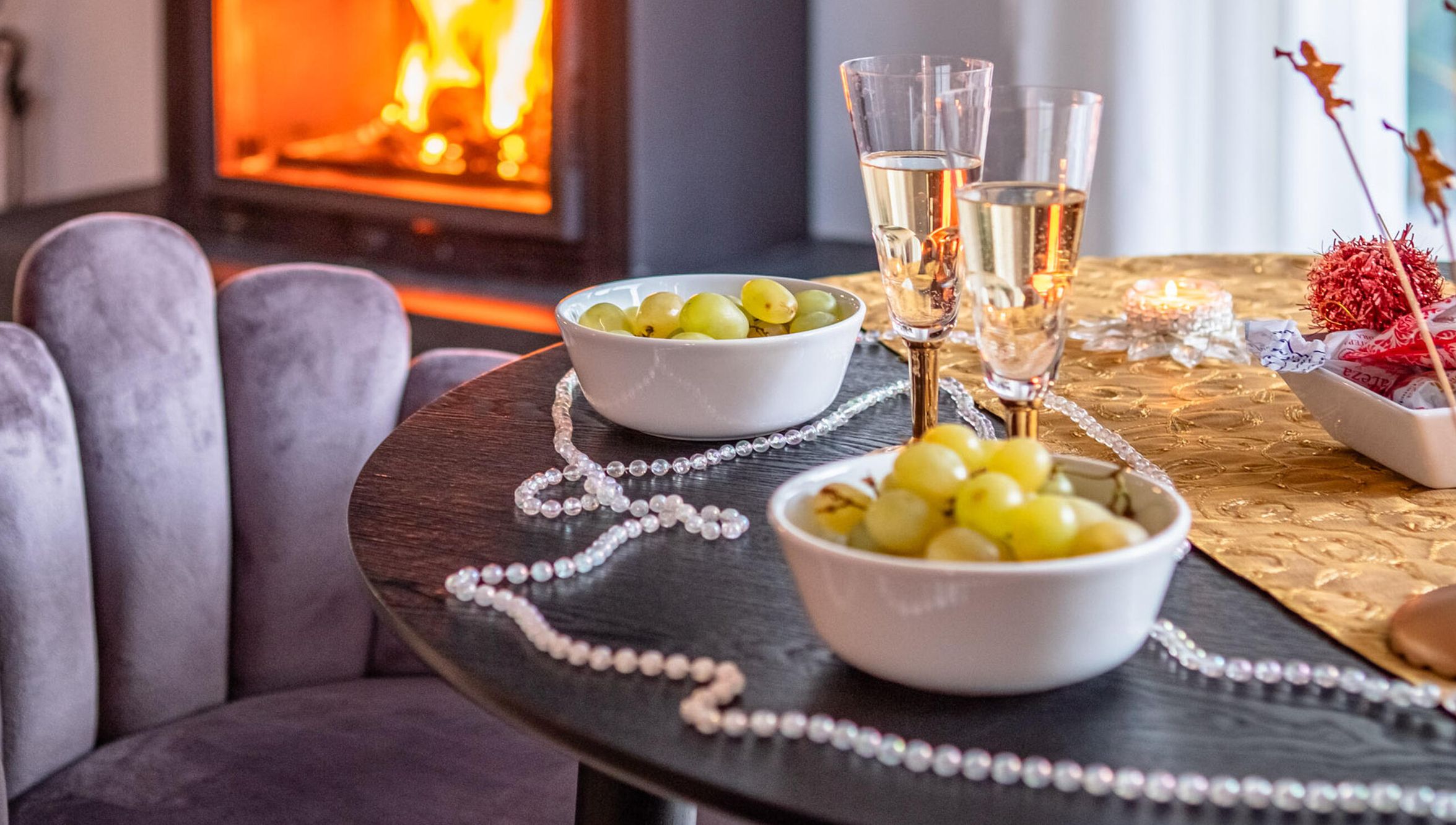 A round table with two bowls of green grapes, a string of pearls, and two glasses of a clear liquid, possibly wine, on a tablecloth with a gold pattern.