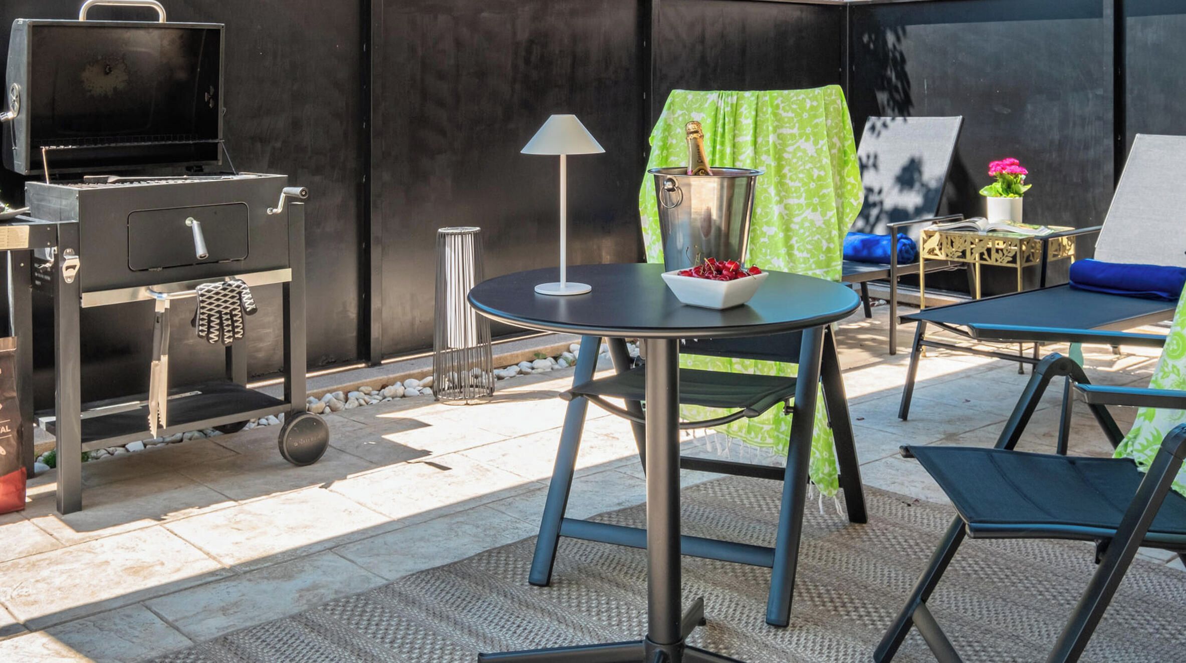 An outdoor patio with a grill, a table with a bowl of red items, a lamp, and a green curtain.