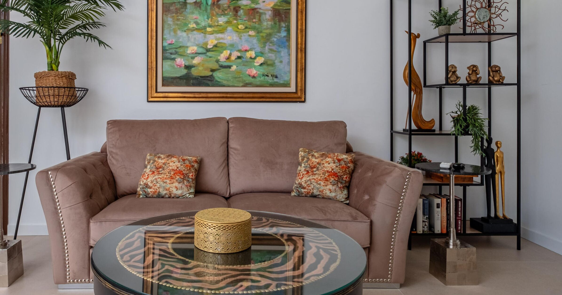 A living room with a brown sofa, two patterned pillows, a round glass coffee table, a framed painting of water lilies, a tall plant in a basket, and a black metal shelf with decorative items.
