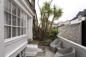 A narrow outdoor space with a white building, a stone wall, and a small garden with plants and wicker furniture. - 18th Century Cottage with Garden, Central Newquay (Cornwall)