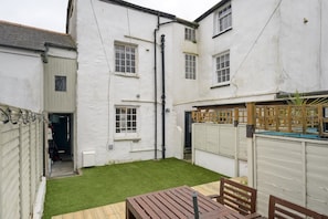 A white building with multiple windows, a small lawn, and a wooden table with chairs. - 18th Century Cottage with Garden, Central Newquay (Cornwall)