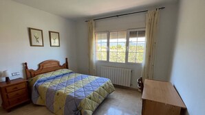 Une chambre à coucher comprenant un lit, une table de chevet en bois, une commode, une fenêtre avec des rideaux et un radiateur.