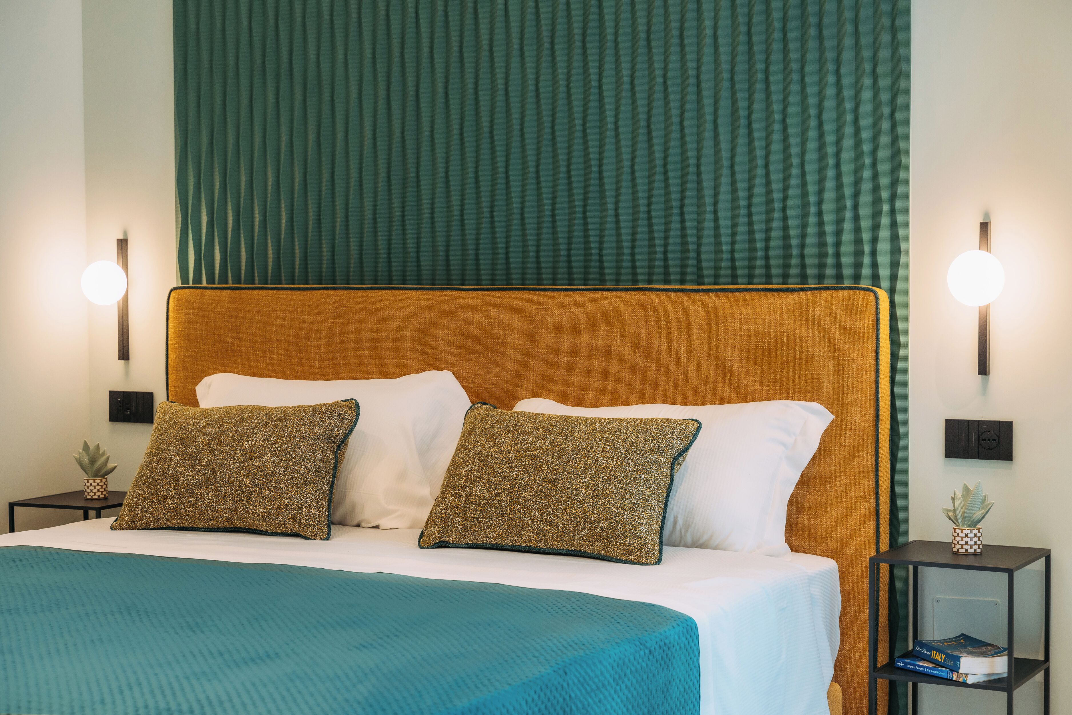 A neatly made bed with a teal bedspread, two pillows, and a wooden headboard. There are two bedside tables with lamps and a small plant on one.