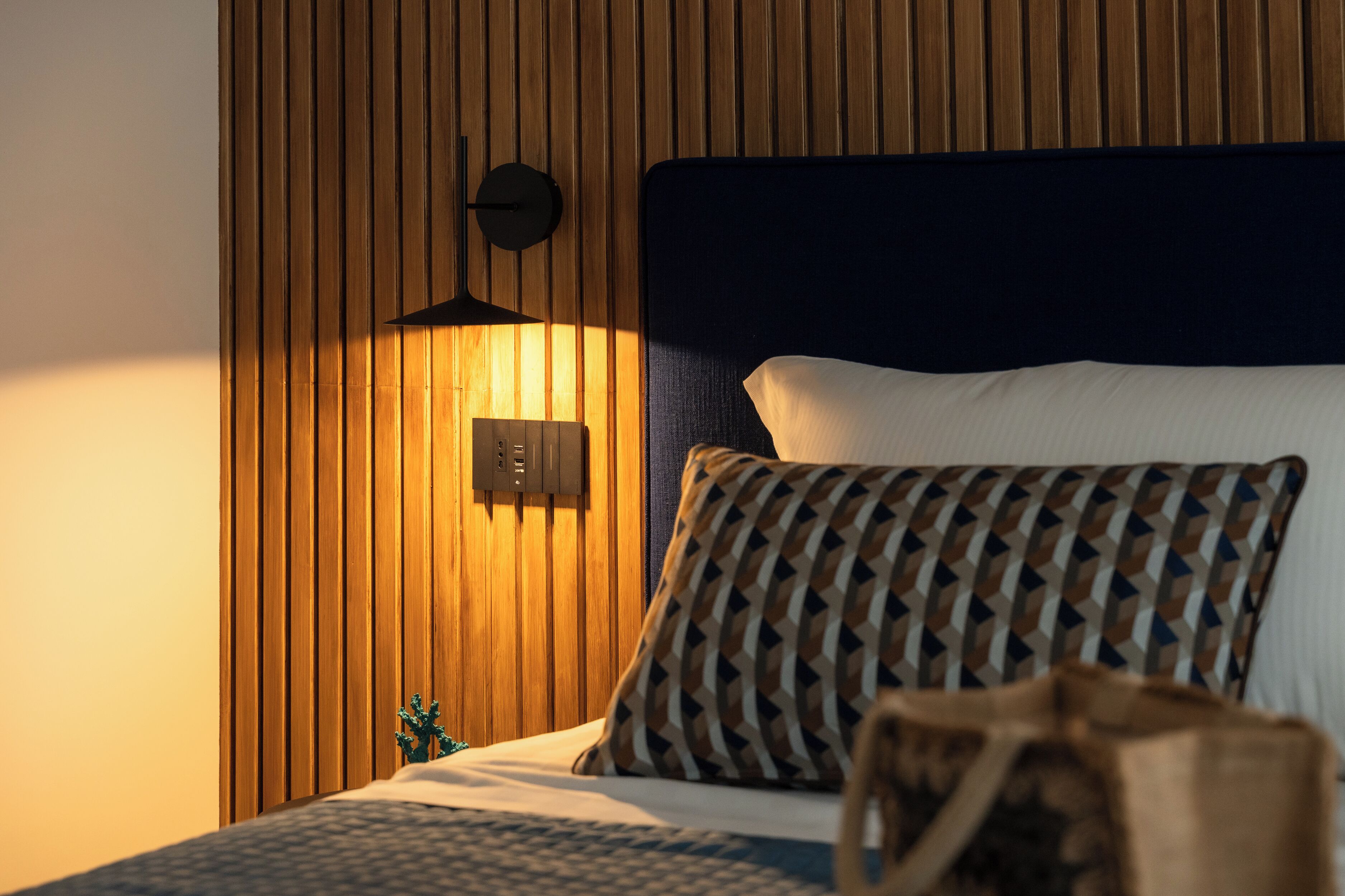 A neatly made bed with patterned pillows, a dark headboard, and a wall-mounted light fixture.