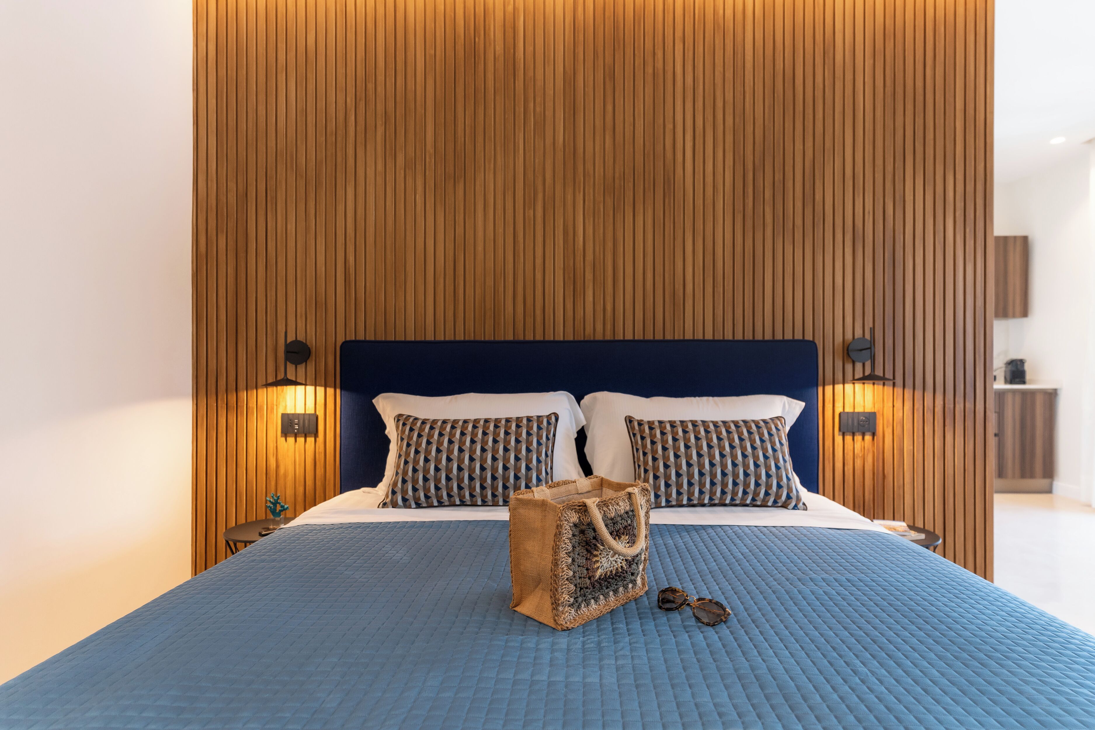 Una cama bien hecha con colcha azul, dos almohadas estampadas y un cabecero de madera. Hay dos lámparas de noche y una mesita con una planta.