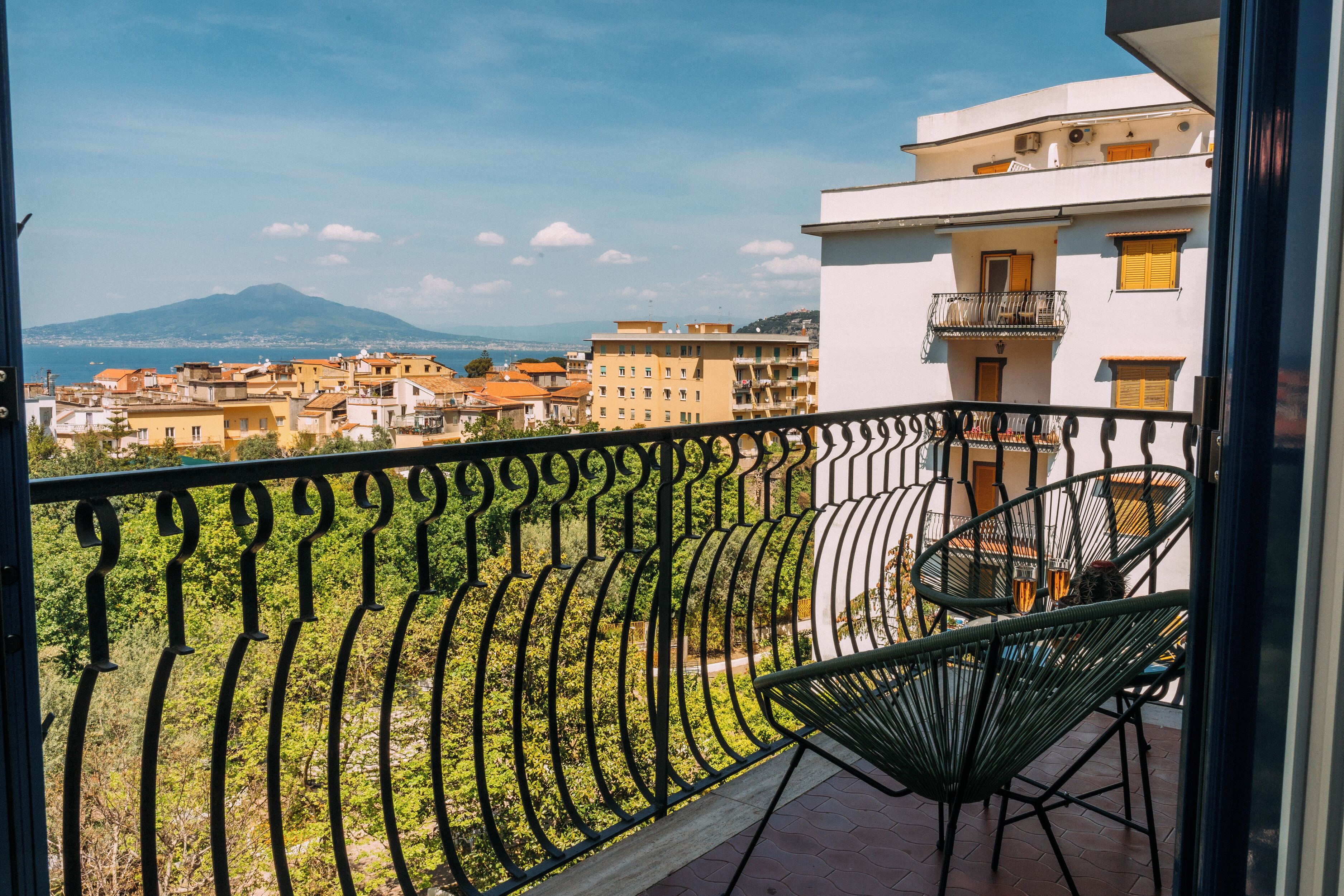 A balcony with a wrought iron railing, a wicker chair, and a view of a cityscape with buildings and a mountain in the distance.
