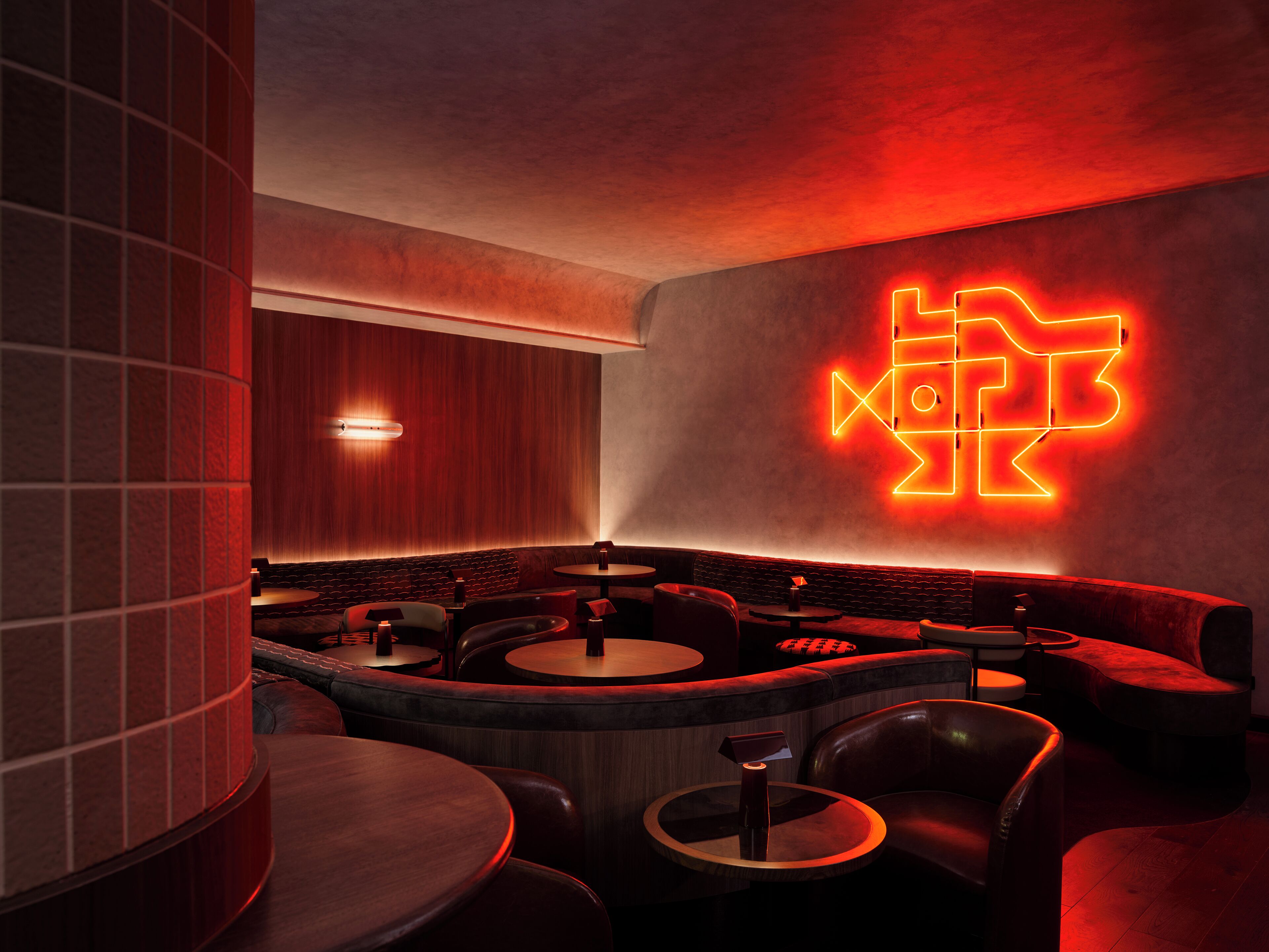 Un área de bar tenuemente iluminada, con mesas redondas y asientos de cuero, un letrero de neón con forma de pez en la pared y una pared de acento de madera.