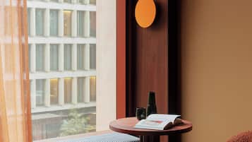 A hotel room with a large window, a round wall-mounted light, a small round table with a book and a bottle, a cushioned bench, and a red blanket.