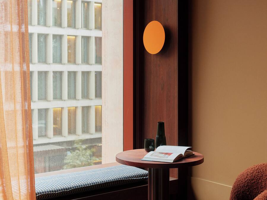 A hotel room with a large window, a round wall-mounted light, a small round table with a book and a bottle, a cushioned bench, and a red blanket.