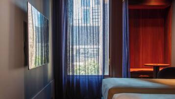 A hotel room with a wooden ceiling, a large window with curtains, a bed, a small table, and a wall-mounted TV.