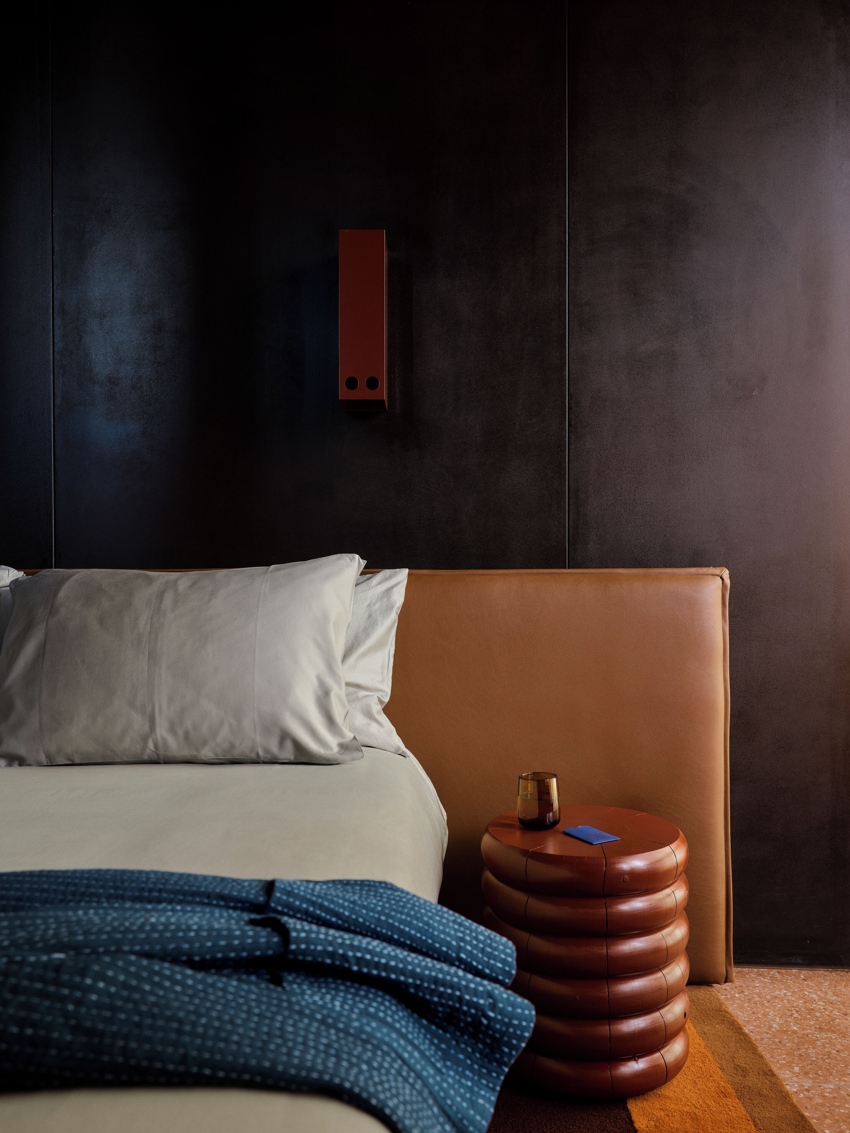 A modern bedroom with a large bed, a headboard, a small round table with a glass, and a wall-mounted light fixture.