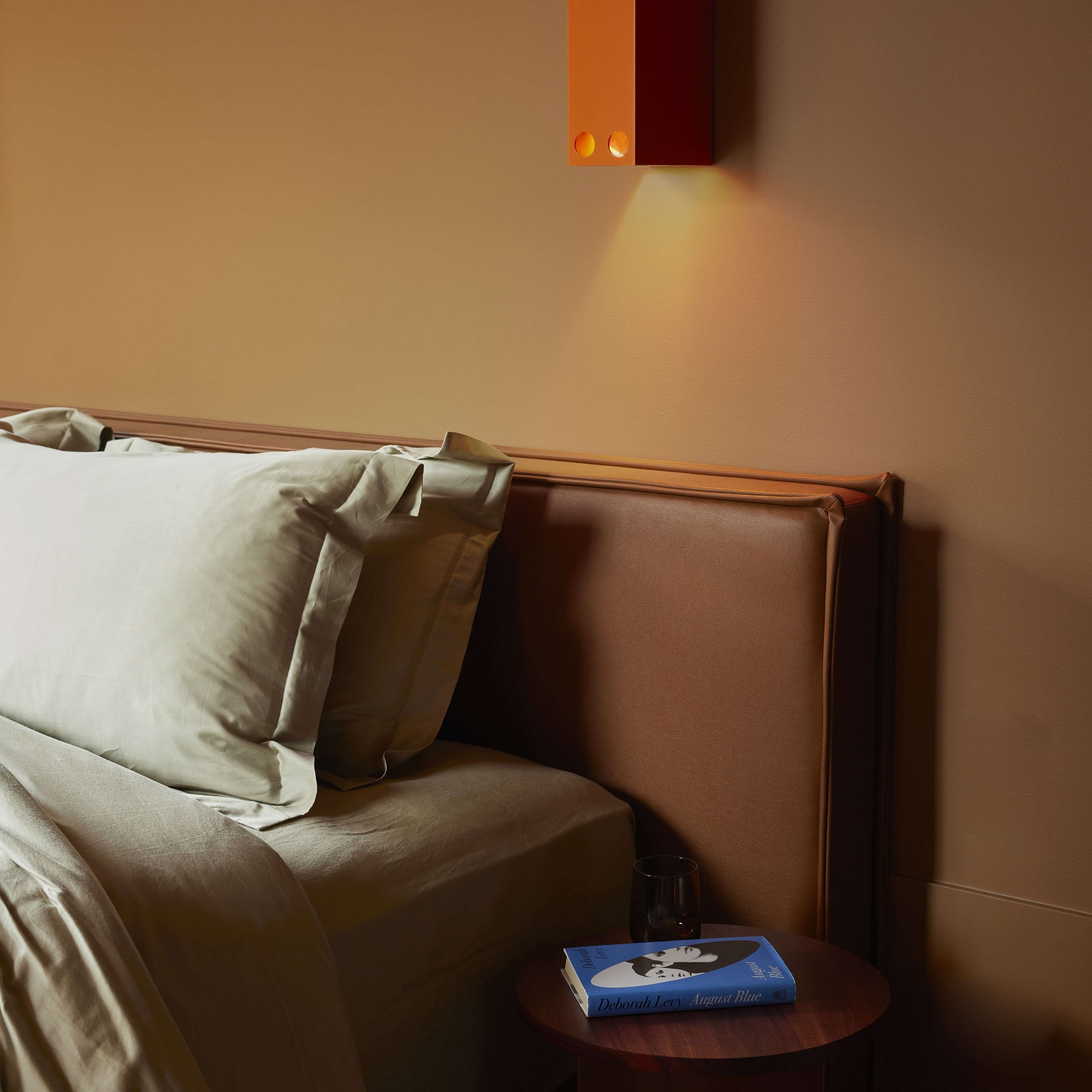 Un dormitorio con una cama, almohadas, una mesita de noche con un libro, y una lámpara fijada a la pared.
