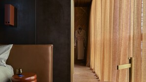 Una habitación de hotel moderna con una cama, un taburete de madera redondo, una bata colgada y vistas a través de una puerta de madera.