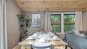 Een eetgedeelte met een houten tafel gedekt voor vier personen, omringd door stoelen, met een grijze bank en een raam met gordijnen op de achtergrond.
