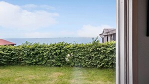 Blick aus einem Fenster auf eine grüne Fläche mit einer Hecke, ein Gebäude mit rotem Dach und ein Gewässer in der Ferne.