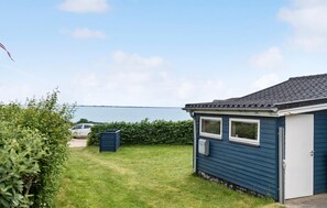 A small, single-story house with a dark blue exterior, a white garage door, and a clear view of the water in the background.