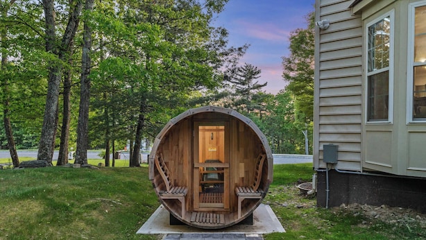 A wooden barrel structure with a door and steps, situated on a paved path in a grassy area with trees in the background.