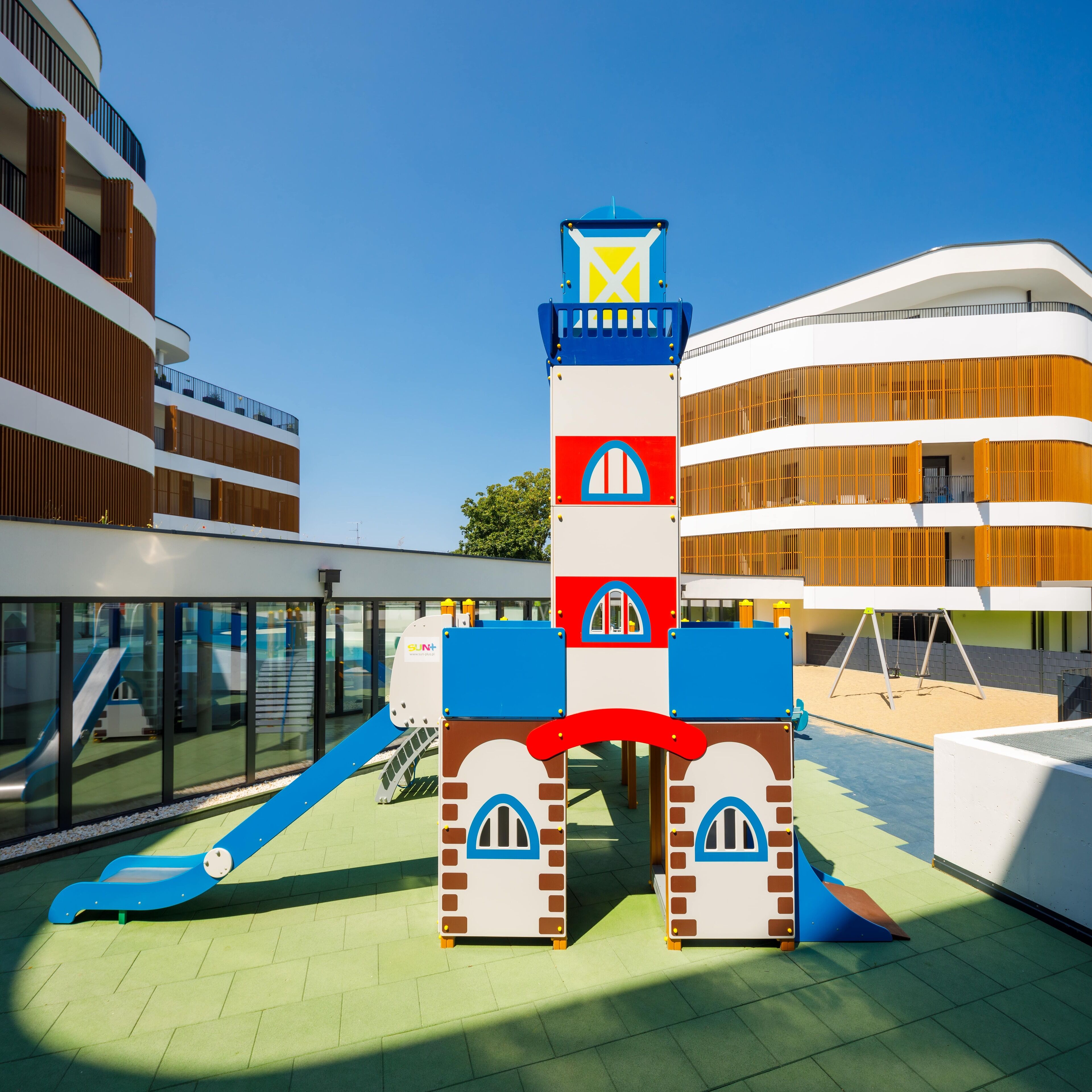 Estructura de juegos infantiles con tobogán y zona de escalada, rodeada de edificios modernos con balcones y una piscina.