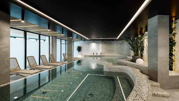 A modern indoor pool with a glass barrier, lounge chairs, and a mosaic-tiled edge.