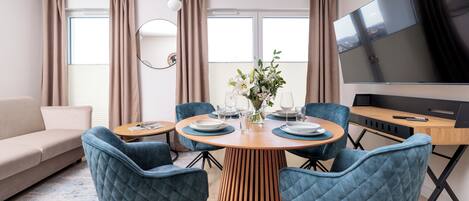 Una sala moderna con una mesa redonda de madera, sillas tapizadas en azul, un sofá color beige, un televisor de pantalla plana y ventanas grandes con cortinas.
