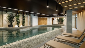 A modern indoor pool area with mosaic-tiled edges, lounge chairs, and potted plants.