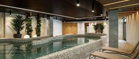 A modern indoor pool area with mosaic-tiled edges, lounge chairs, and potted plants.