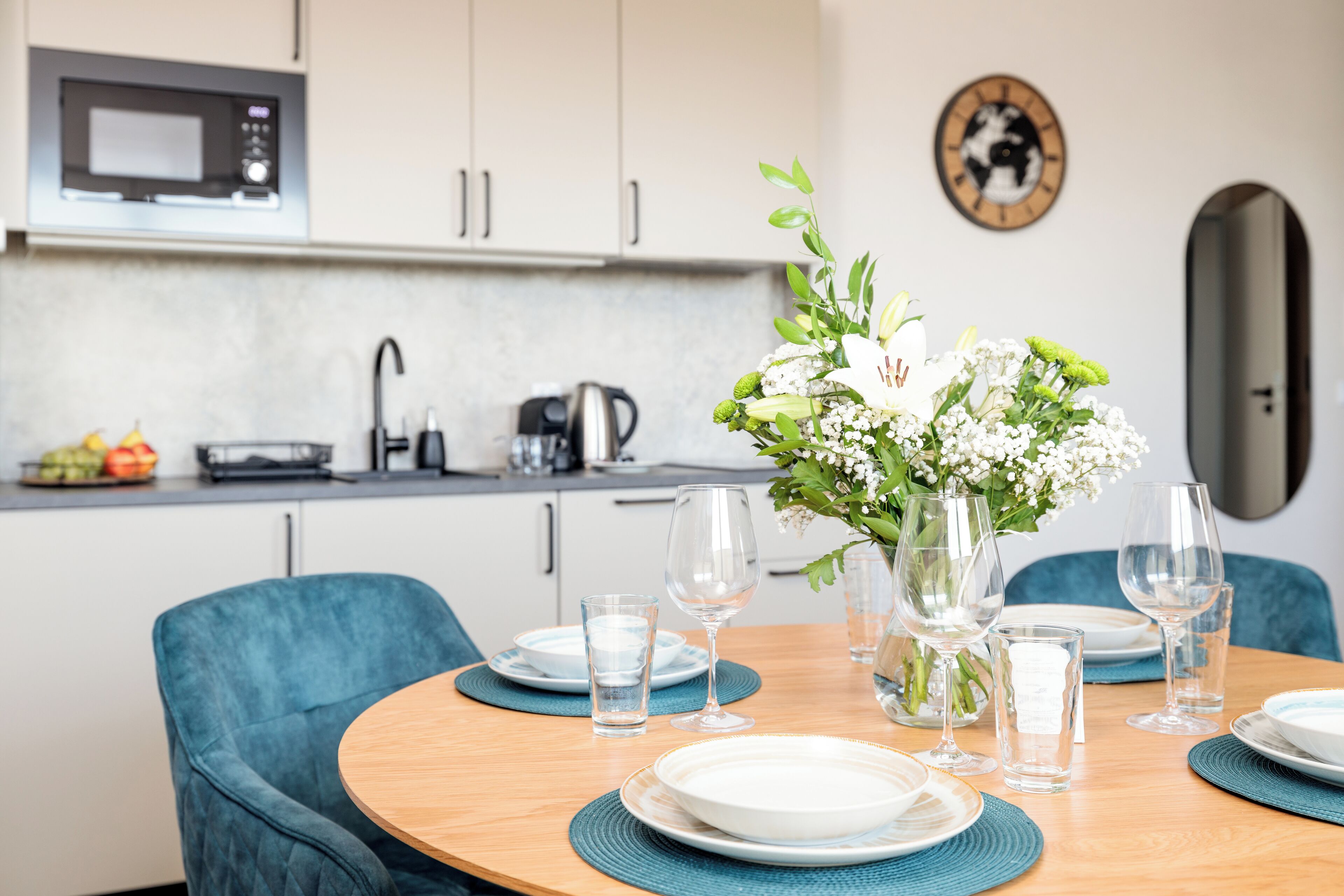A modern kitchen with white cabinets, a microwave, and a sink. There is a dining table set with plates, glasses, and a vase with flowers.