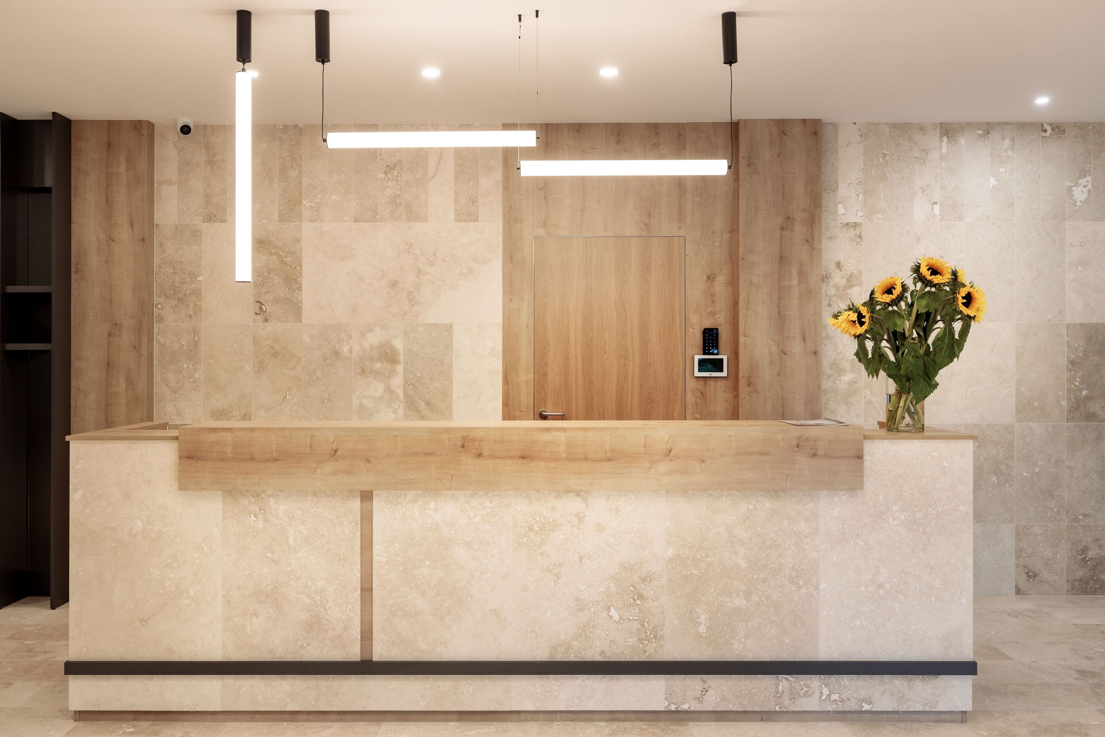 A reception desk with a wooden counter, a wall-mounted control panel, and a vase with sunflowers.
