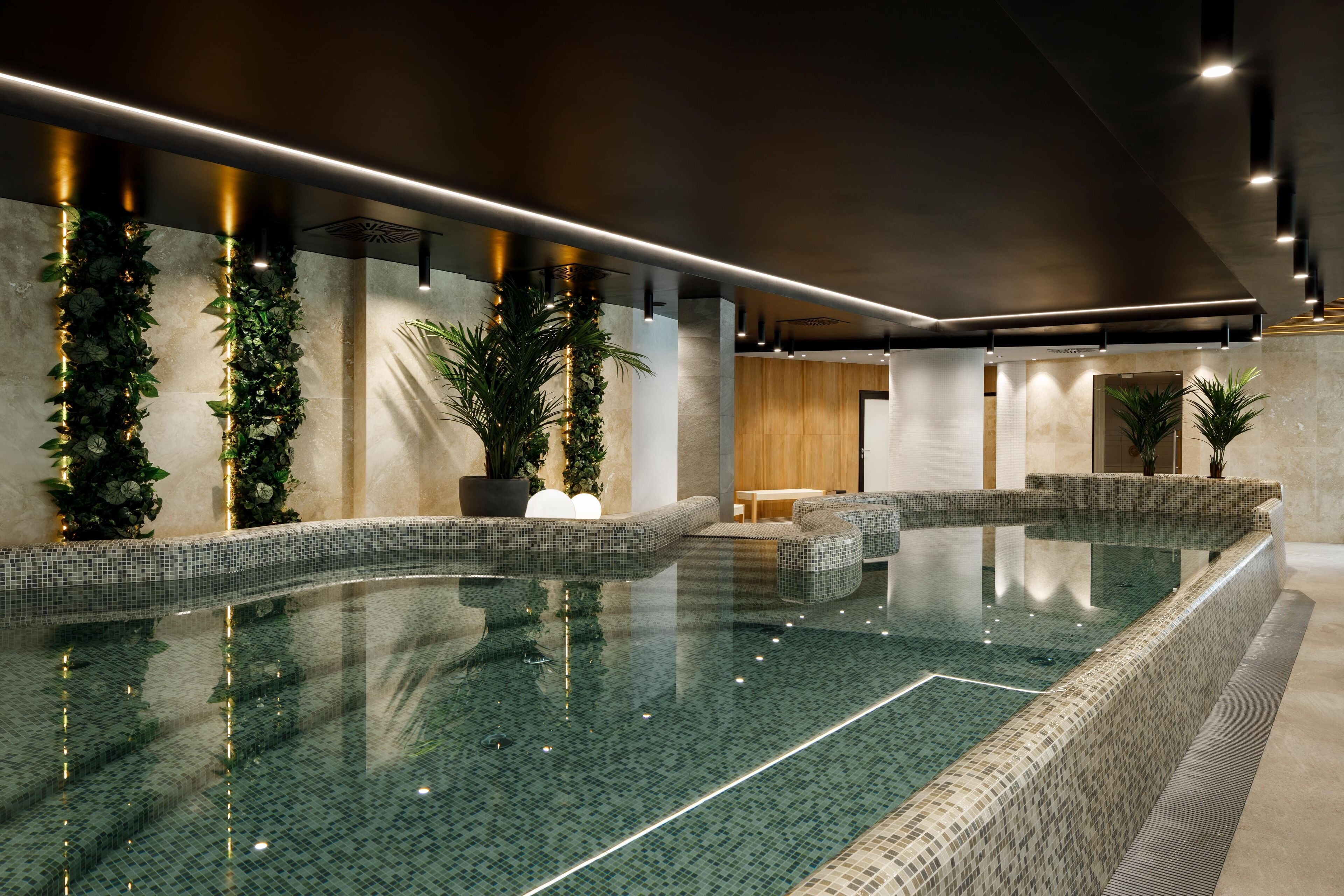 A modern indoor swimming pool with a tiled floor and walls, surrounded by potted plants and a wooden reception area.