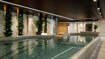 A modern indoor swimming pool with a tiled floor and walls, surrounded by potted plants and a wooden reception area.