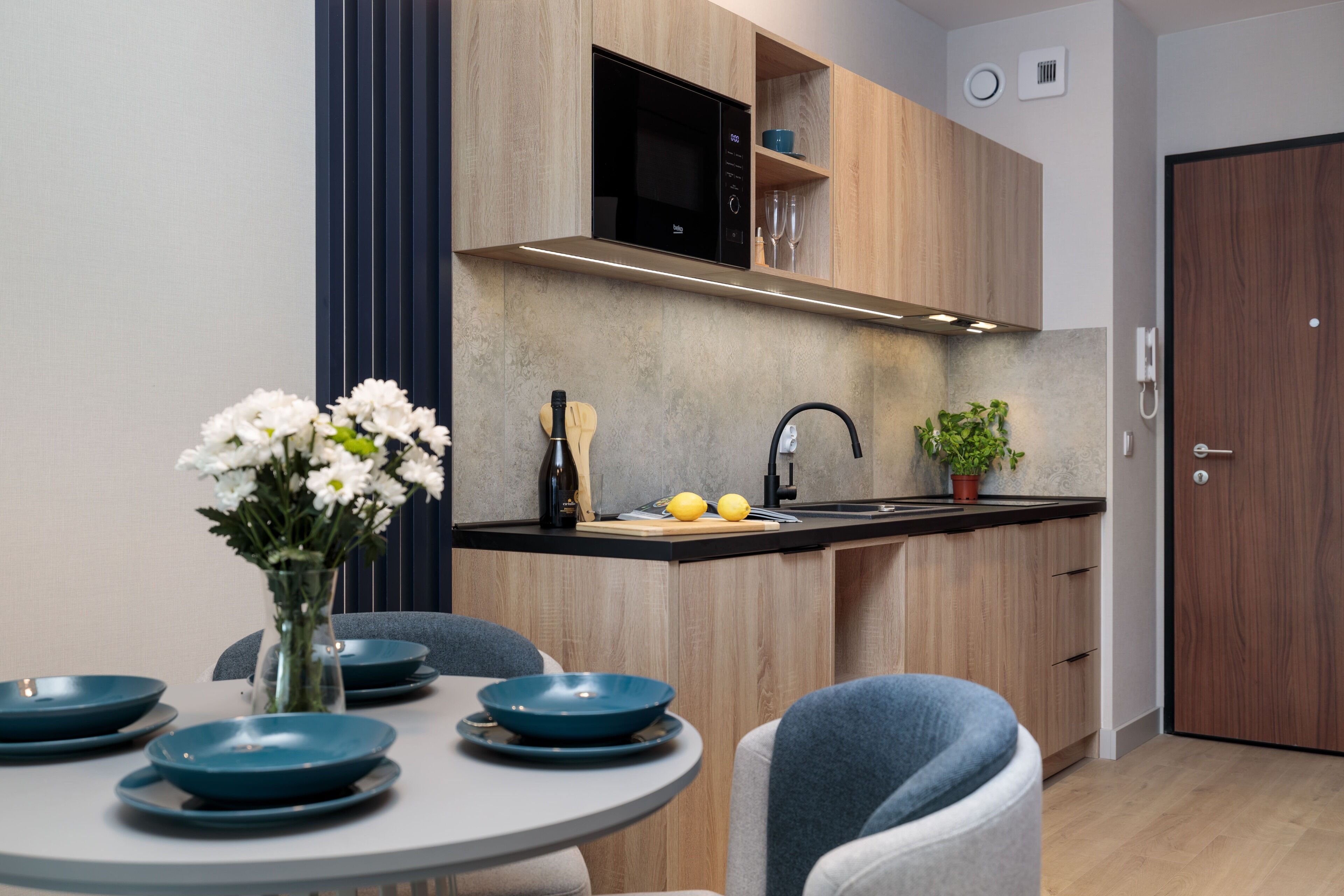 A modern kitchen with wooden cabinets, a black microwave, and a sink. There is a dining table with blue plates and a vase of flowers.