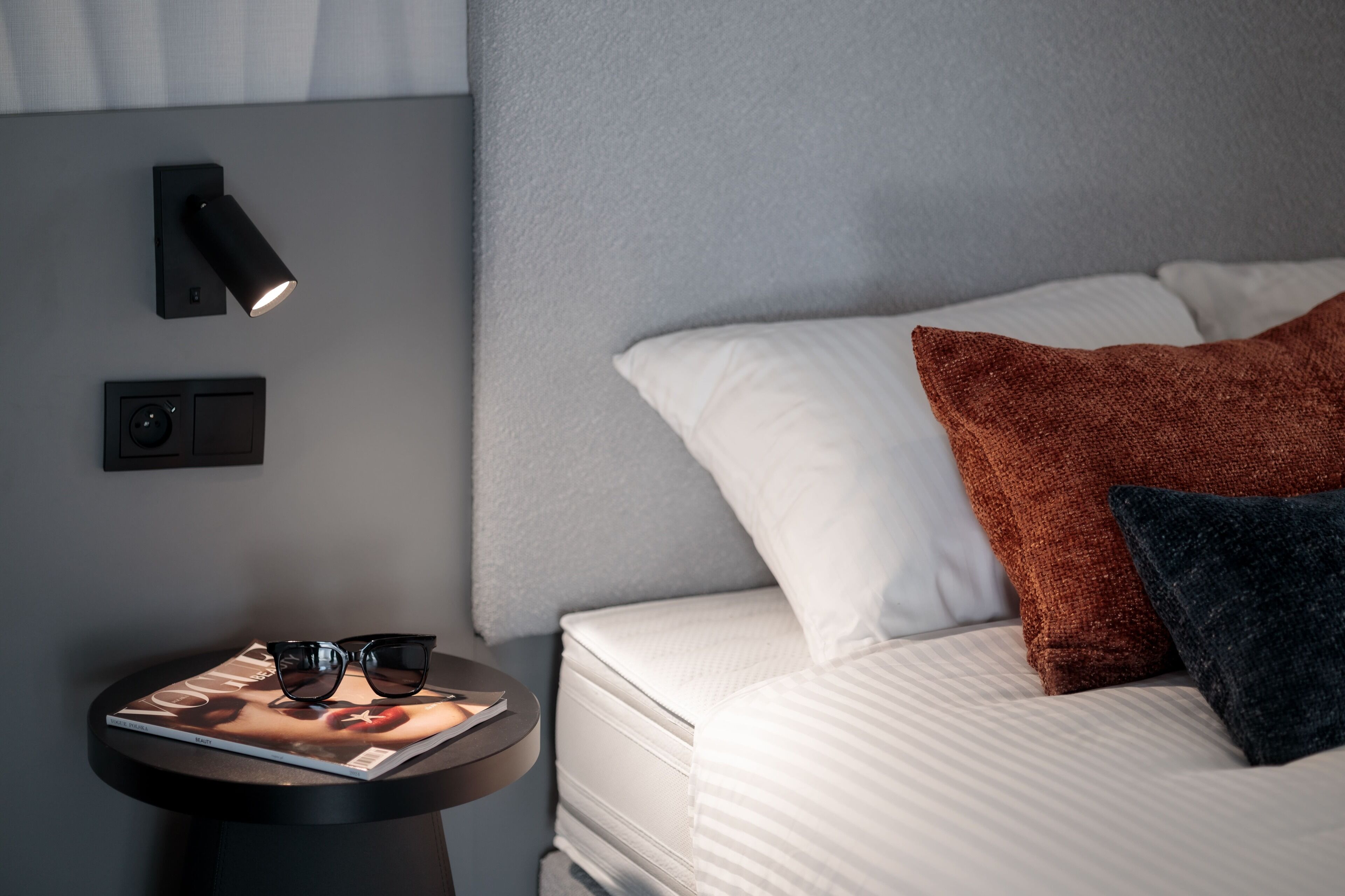 A neatly made bed with white and brown pillows, a small table with a magazine and glasses, and a wall-mounted light fixture.