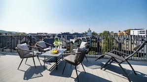A rooftop terrace with outdoor furniture, a table with a fruit basket, and a view of buildings and a tower in the distance.