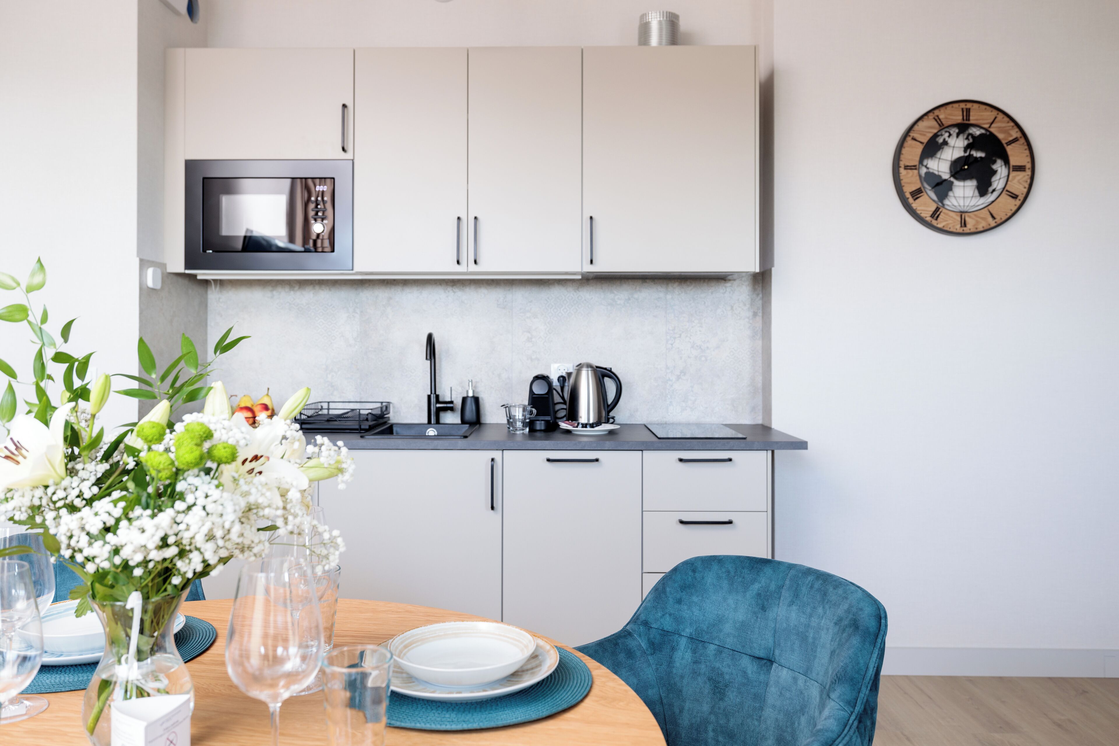 A modern kitchen with white cabinets, a microwave, and a kettle. There is a dining table with plates and glasses, and a vase of flowers.