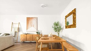 Een eetgedeelte met een houten tafel en bankjes, een witte muur met een inlijsting spiegel, en een ladder tegen de muur.