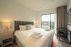 A hotel room with a large bed, two bedside lamps, a desk with a chair, and a view of the city through a large window. - Beautiful, modern, light flat in Shoreditch (a) (Greater London)