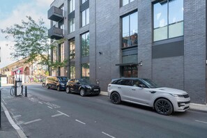 A modern building with a brick facade, large windows, and a parking area with two SUVs. - Beautiful, modern, light flat in Shoreditch (a) (Greater London)